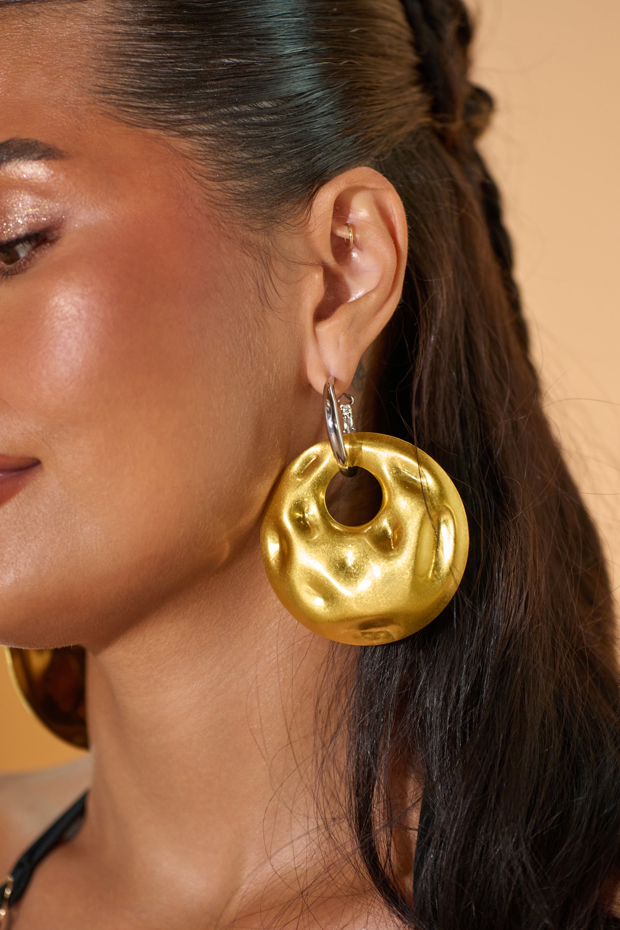 A close-up shows a woman's ear adorned with the LEMME IN EARRING—a large, round, hammered disc earring featuring a circular cutout at the top—standing out against her warm tan skin and dark hair swept back. 0