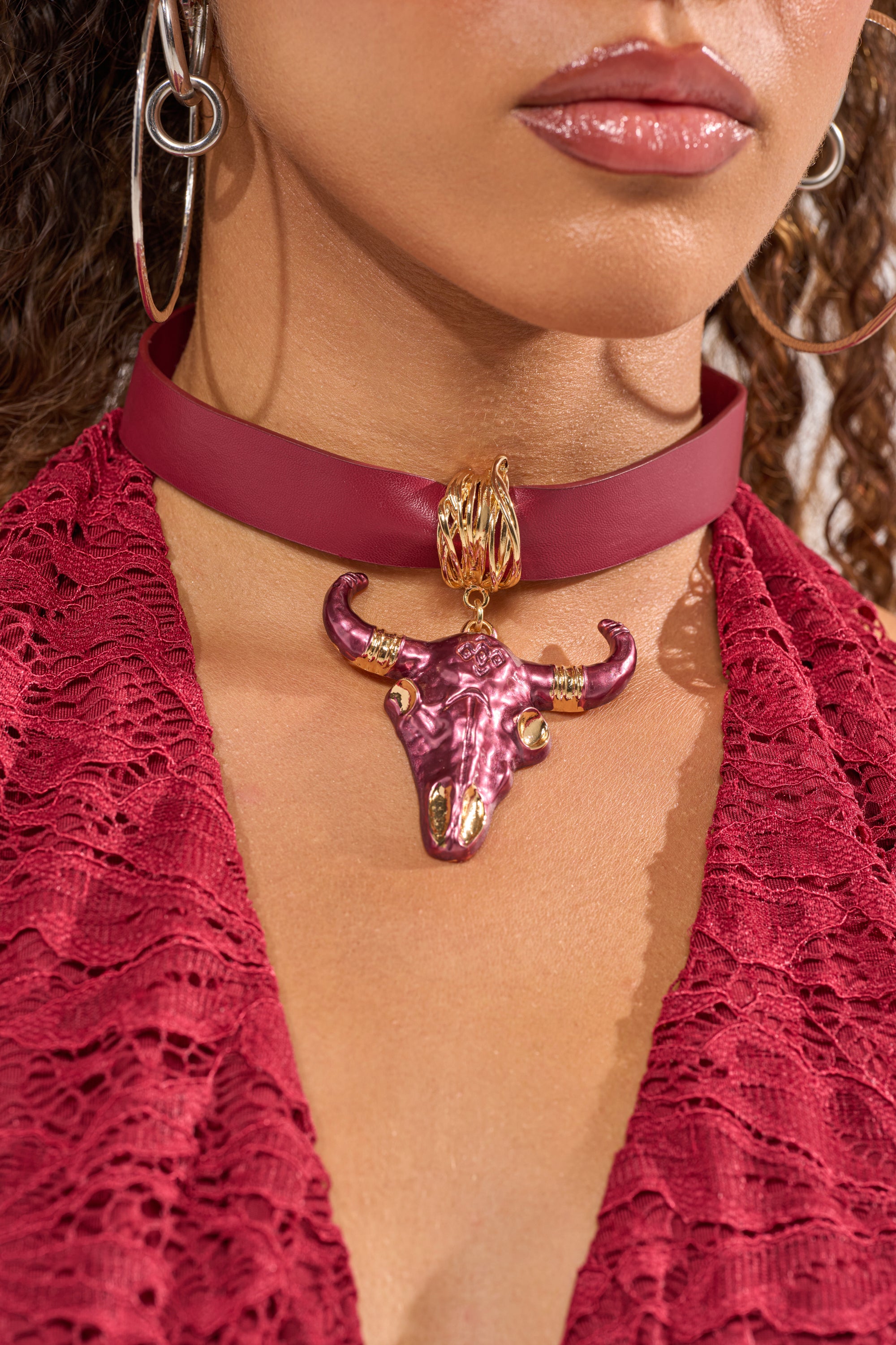 A woman wears a deep red lace top, large hoop earrings, and the RED HOT NECKLACE, which features a gold and maroon bull skull pendant. Only her lower face and neckline are visible. 1