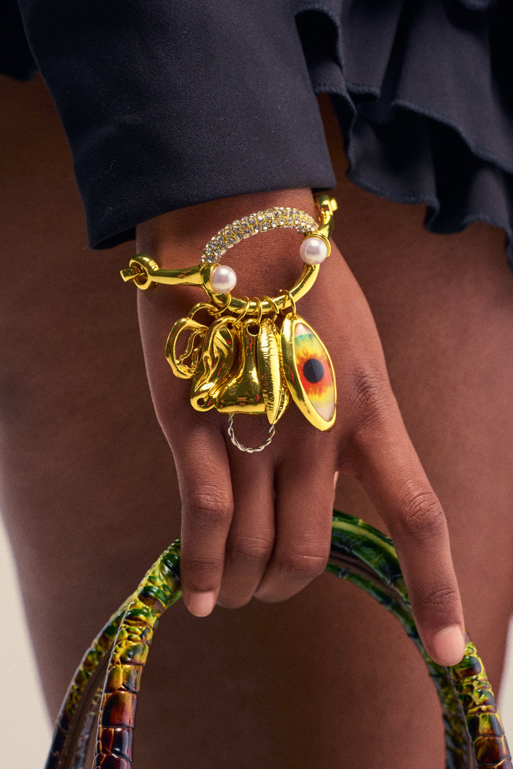 A hand holds a textured green purse, wearing the ONE EYE OPEN BRACELET—a gold rhinestone bracelet adorned with charms, pearls, and sparkling details. The person wears a black ruffled garment.