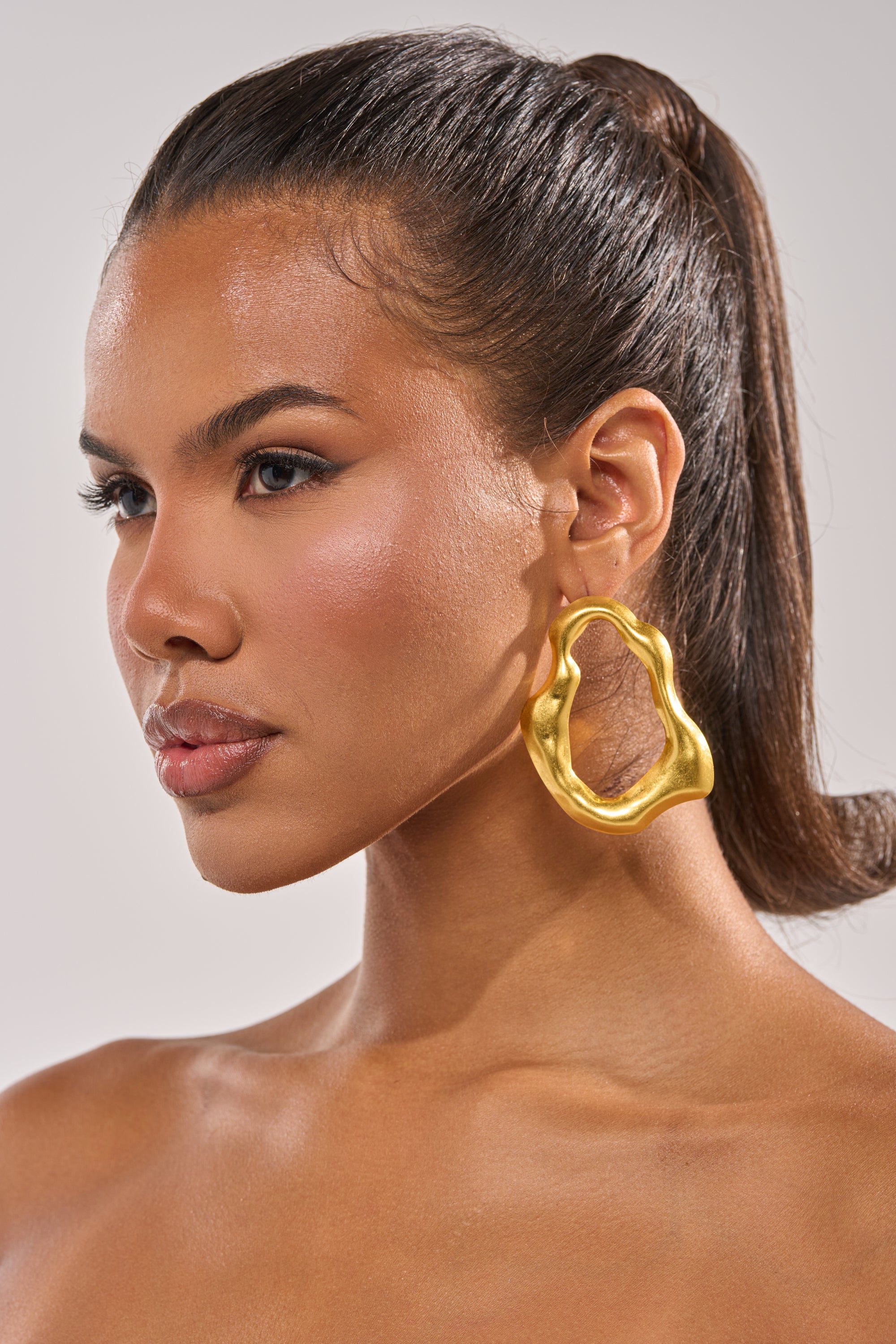 A woman with smooth, glowing skin and straight, dark hair pulled back models the sculptural gold TEMPTED EARRING. She is shown in profile against a neutral background. 1