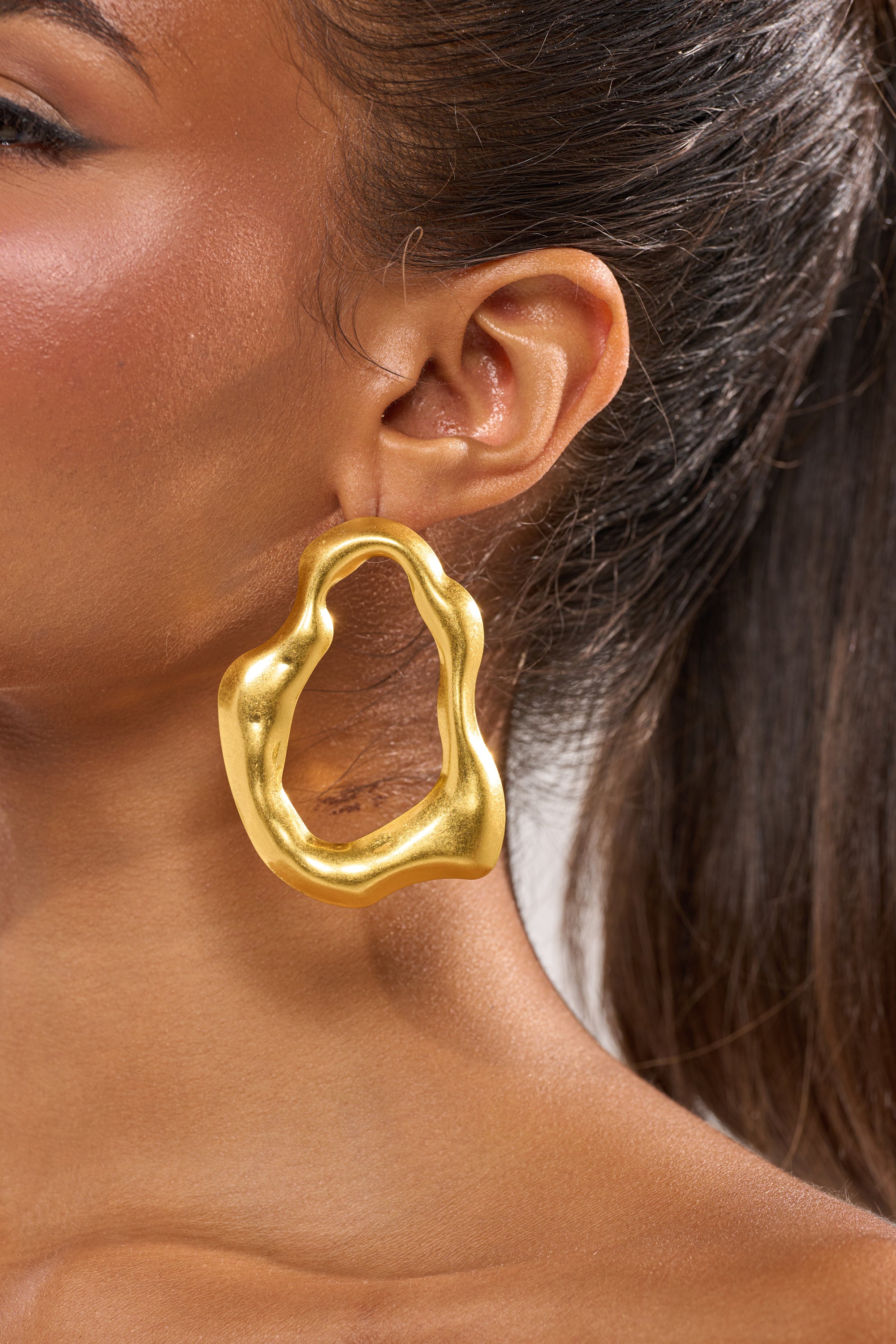 Close-up of a woman's ear adorned with the TEMPTED EARRING, a large sculptural gold drop style. Her hair is pulled back to reveal the striking accessory, and her skin glows with a smooth radiance. 2