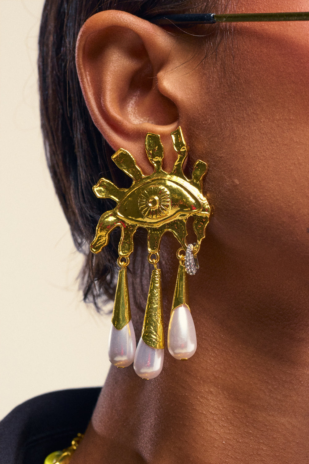A close-up of a person wearing the BE MINE EARRING, a large gold-tone earring shaped like an eye with radiating lines and three dangling faux pearl drops.