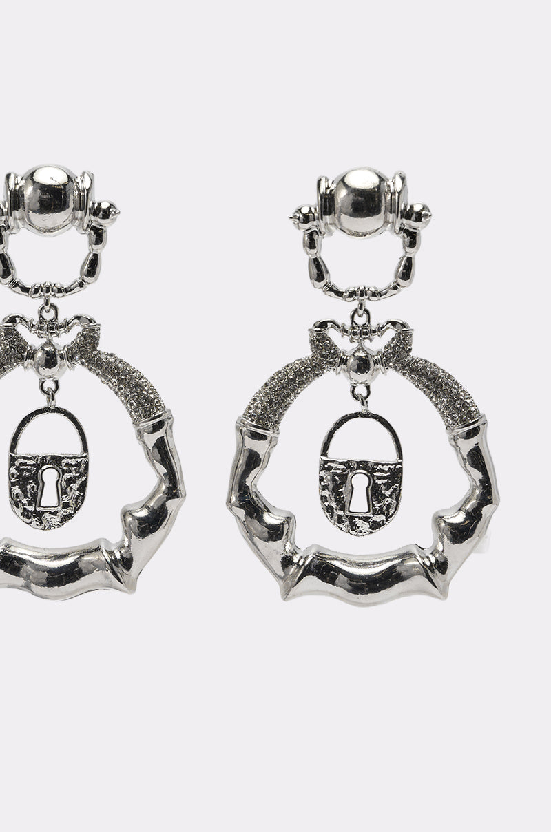 Close-up of the FLEW OUT EARRING: silver statement drops with a textured circular hoop, a bow at the top, and a lock charm featuring a keyhole at the center. Set against a plain white background. 2