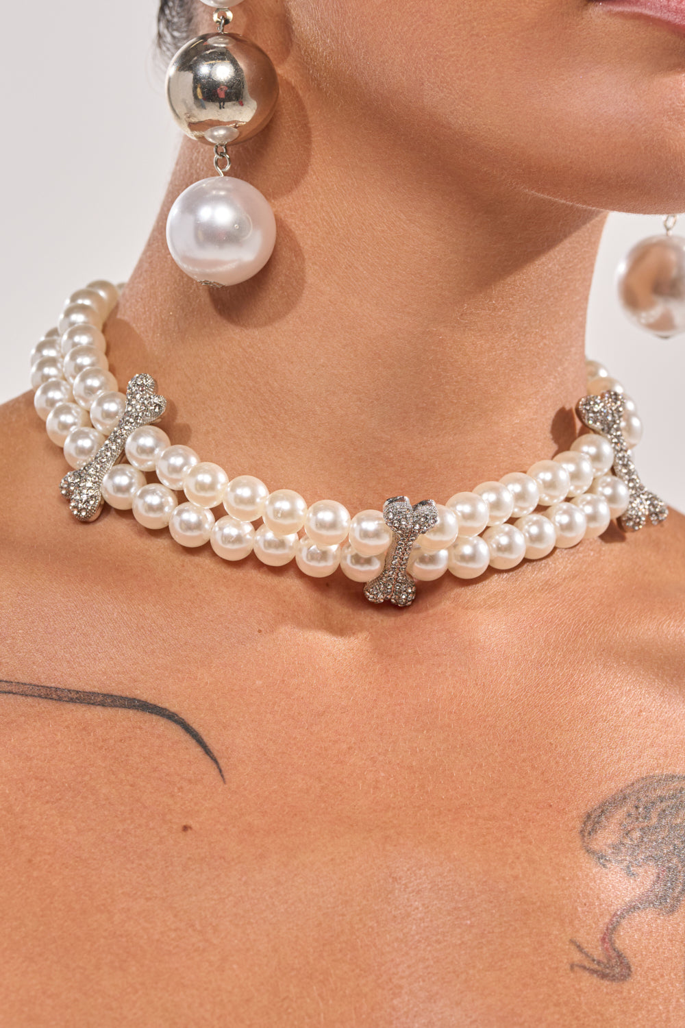 Close-up of a person wearing the ALIA PEARL NECKLACE featuring layered pearls and rhinestone bone charms, paired with large pearl drop earrings; two tattoos are partially visible on their upper chest and shoulder. 1