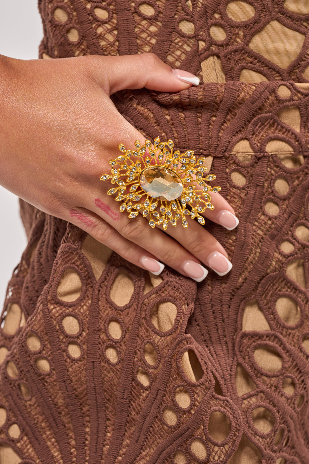 A close-up of light pink manicured nails wearing the TAKE ME OUT RING, a bold gold cocktail piece with a sunburst design and yellow gemstone, resting on an intricately patterned brown lace dress. 1