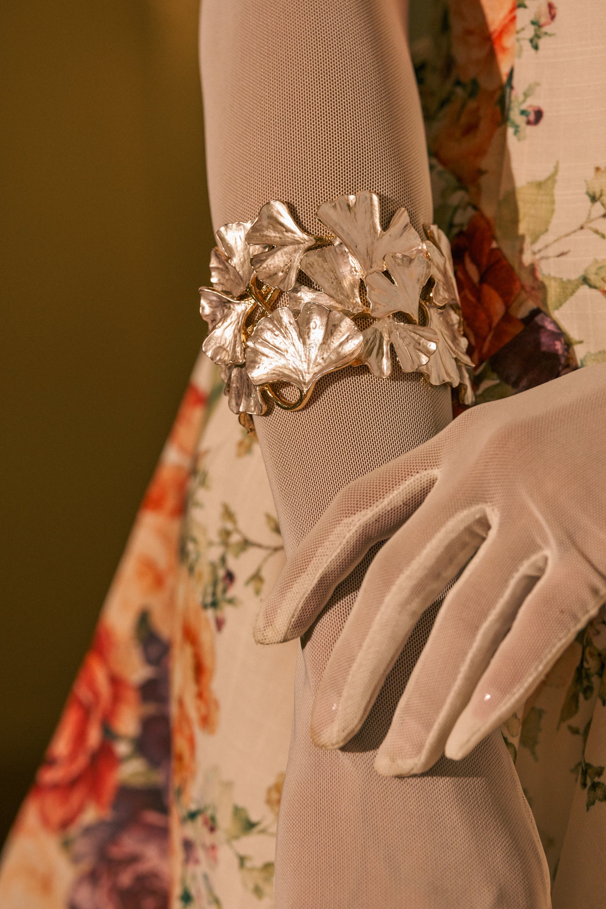 A close-up of a person wearing sheer white gloves and a floral dress, with the arm gently bent and the hand resting on the dress, showcases the IN BLOOM BRACELET—a pearlescent floral cuff adorned with delicate detailing—making this elegant accessory an ideal choice for a bridal bracelet.