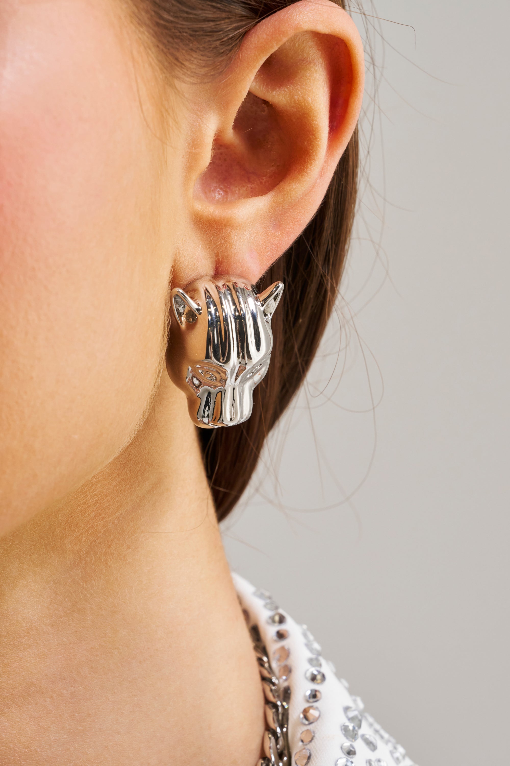 Close-up of a woman's ear wearing the BITE ME EARRING—metallic silver studs shaped like a cat's face. Her straight brown hair and white top with metallic accents highlight her bold statement style.