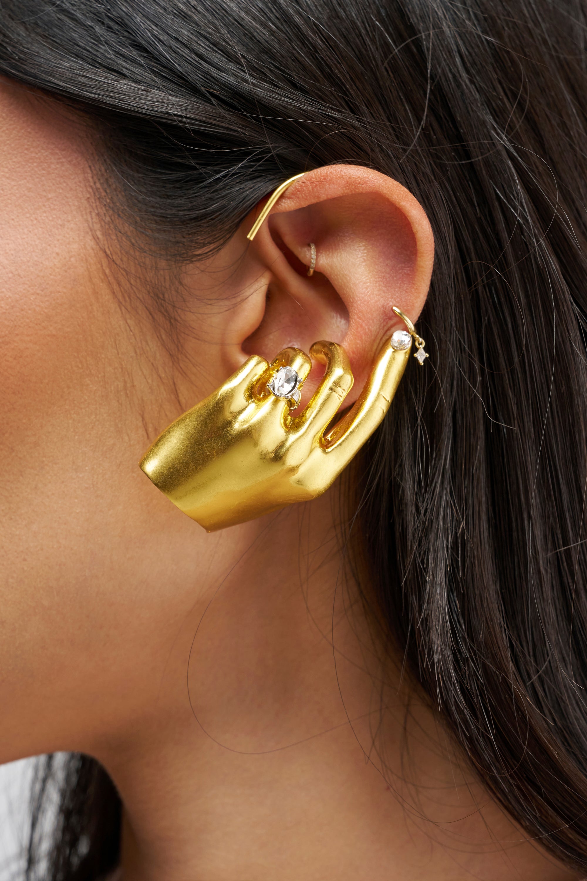 Showcased in a close-up, the ear is adorned with the FIRST CHOICE STATEMENT EARRING CUFF IN GOLD—a bold, hand-shaped gold ear cuff that gently clasps the ear. Paired with several smaller hoop and stud earrings, this unique piece stands out as striking festival jewelry, ideal for making a memorable impression.