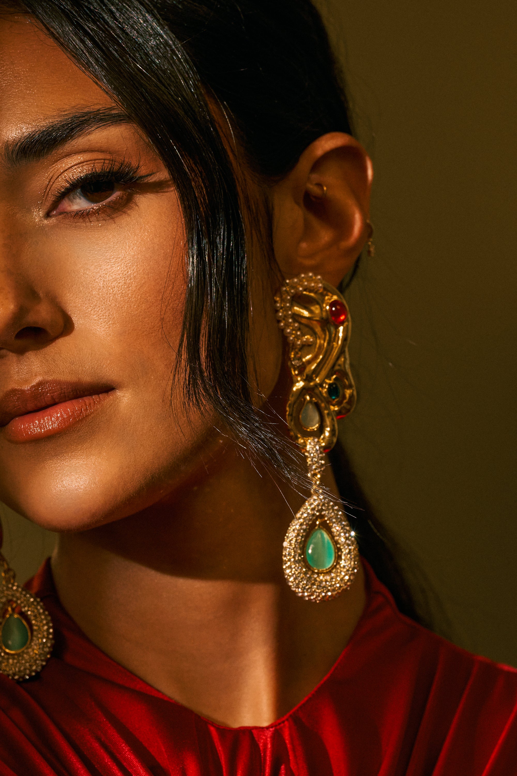 A close-up of a woman wearing the ornate gold AN EARFUL DROP EARRINGS with green stones, dressed in shiny red fabric. Only half of her face is visible, highlighting her eye, lips, and the striking earring against a dark background.