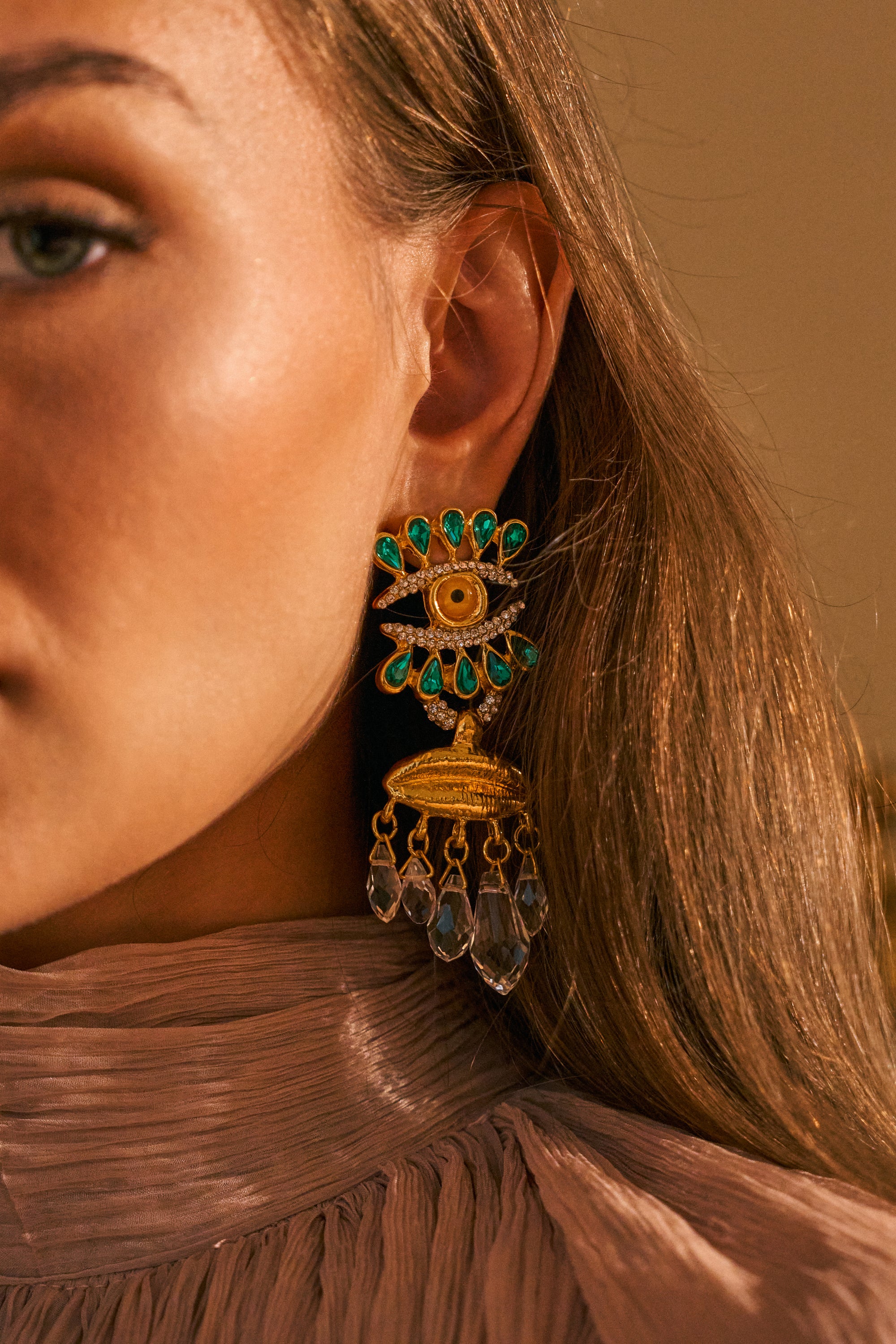 A close-up of a woman’s face shows her wearing the EYE SEE YOU DROP EARRINGS—large gold earrings featuring an eye motif with green stones, rhinestone accents, and dangling crystals. She has long brown hair and wears a pleated, high-neck beige top.