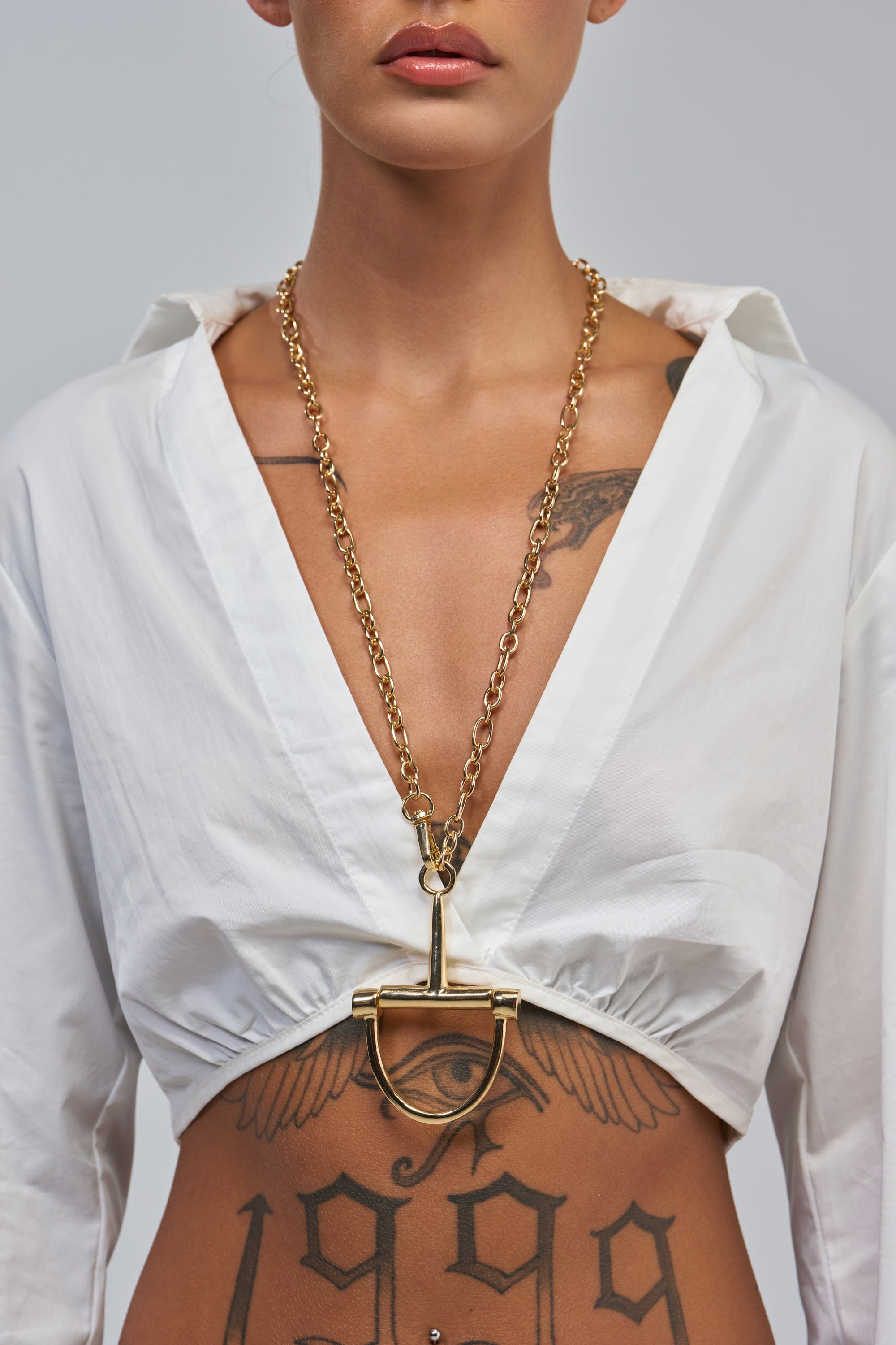 A person pairs a white deep V-neck top with the GOLD ANCHOR Y-NECK and visible “1999” and wing tattoos, creating a bold, layered look.