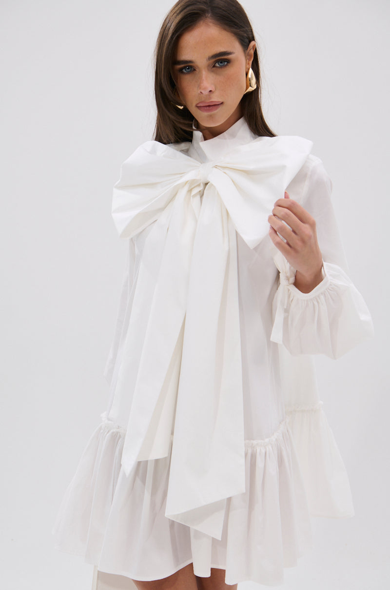Wearing the TAKE A BOW POPLIN MINI DRESS with puff sleeves and a bold white bow at the neckline, a woman stands against a plain light background, her long brown hair framing her face as she looks at the camera.