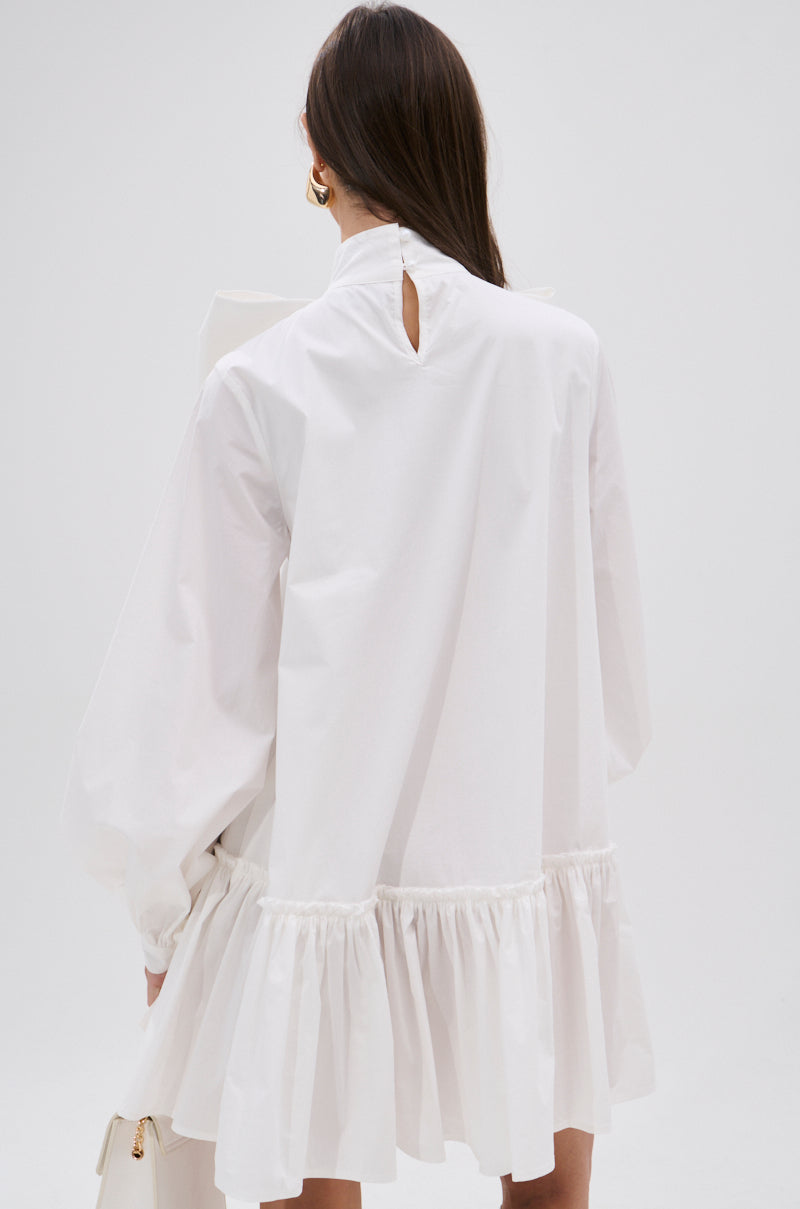 A person with long brown hair, seen from behind, wears the TAKE A BOW POPLIN MINI DRESS featuring gathered ruffles at the hem, puff sleeves, and a small buttoned keyhole on the back. The background is plain and light-colored.