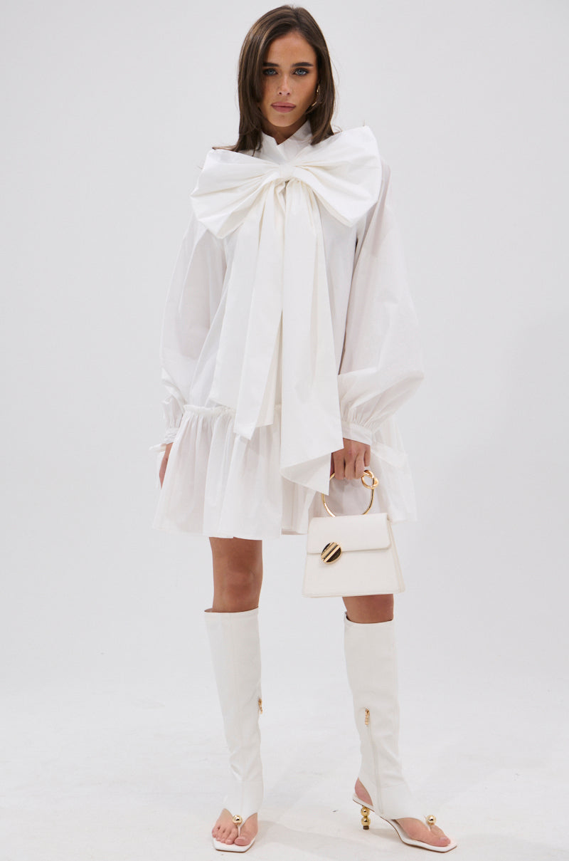 A woman poses against a white background in the TAKE A BOW POPLIN MINI DRESS, which features an oversized bow at the neck. She pairs it with white knee-high heeled boots and carries a small white handbag with a gold handle.