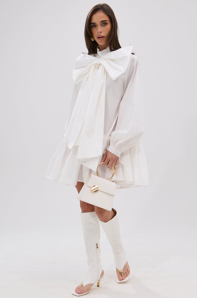 A woman wears the TAKE A BOW POPLIN MINI DRESS, featuring an oversized bow at the collar, paired with a small white handbag and knee-high white heeled boots, as she stands against a plain background.
