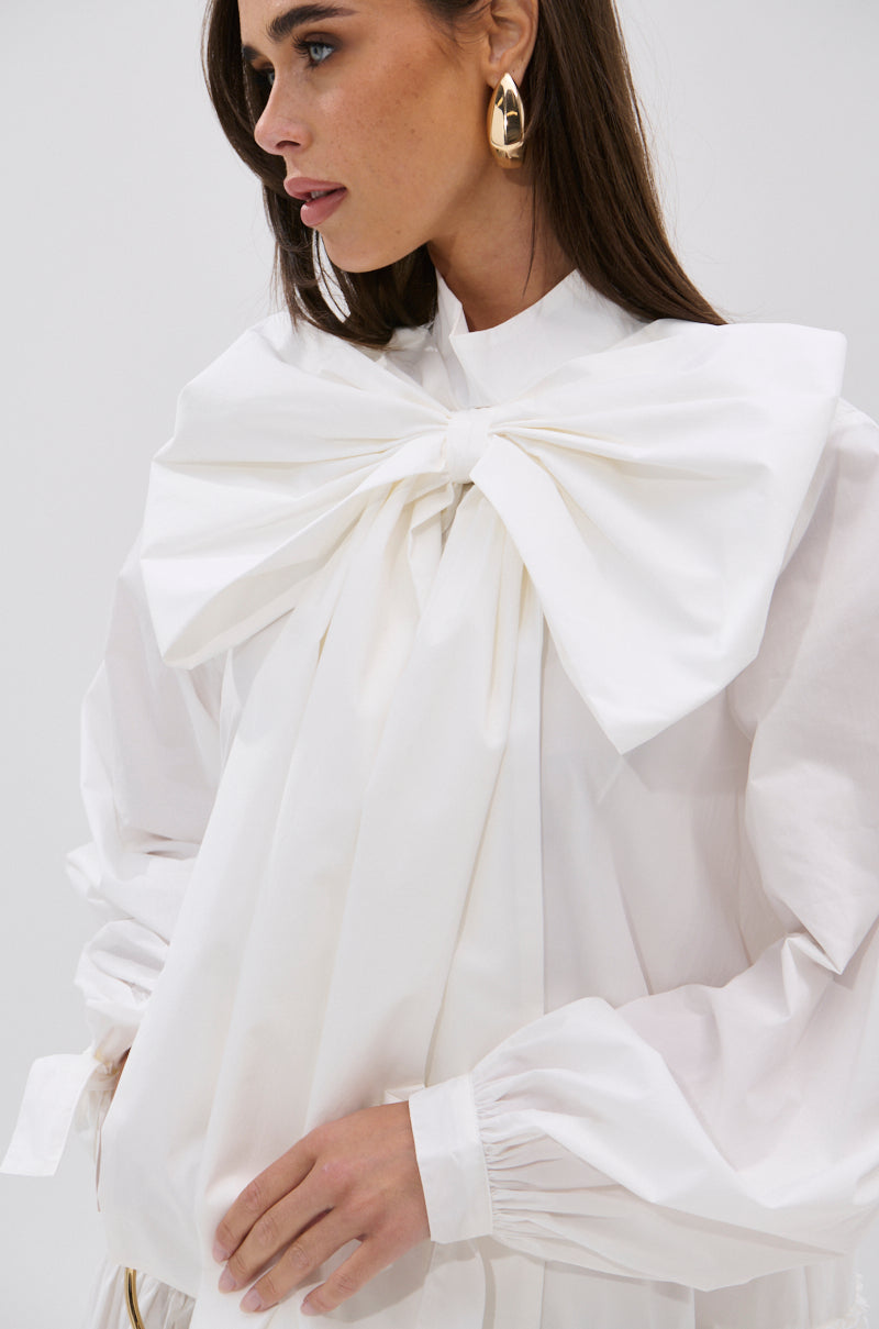A woman with long brown hair and gold statement earrings wears the TAKE A BOW POPLIN MINI DRESS, showcasing puff sleeves and an oversized bow at the collar. She stands against a plain white background with a neutral expression.