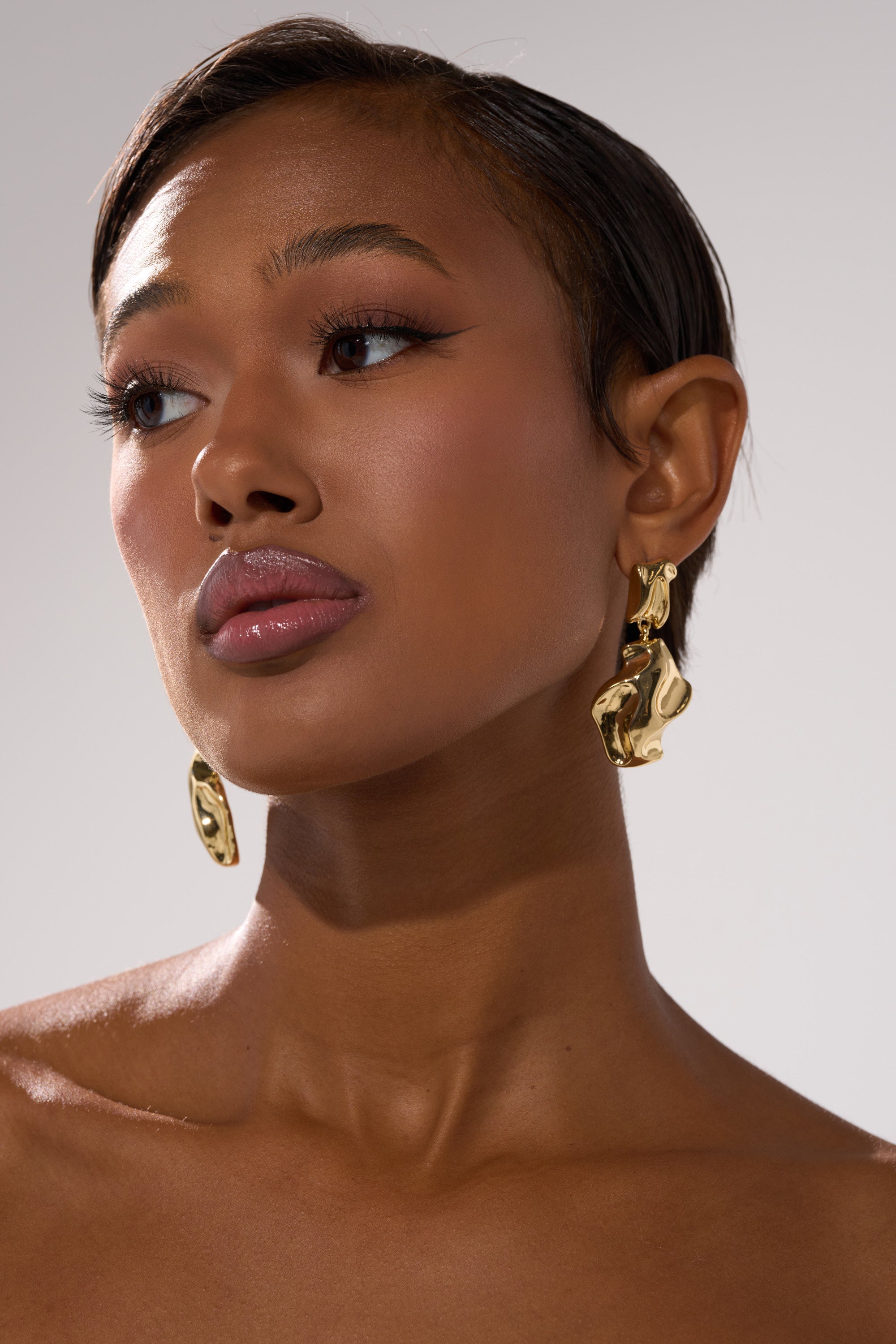 A woman with smooth skin and short, slicked-back hair looks upward, showcasing flawless makeup and glossy lips while wearing bold LEAVE ME ALONE EARRING statement earrings against a neutral background.