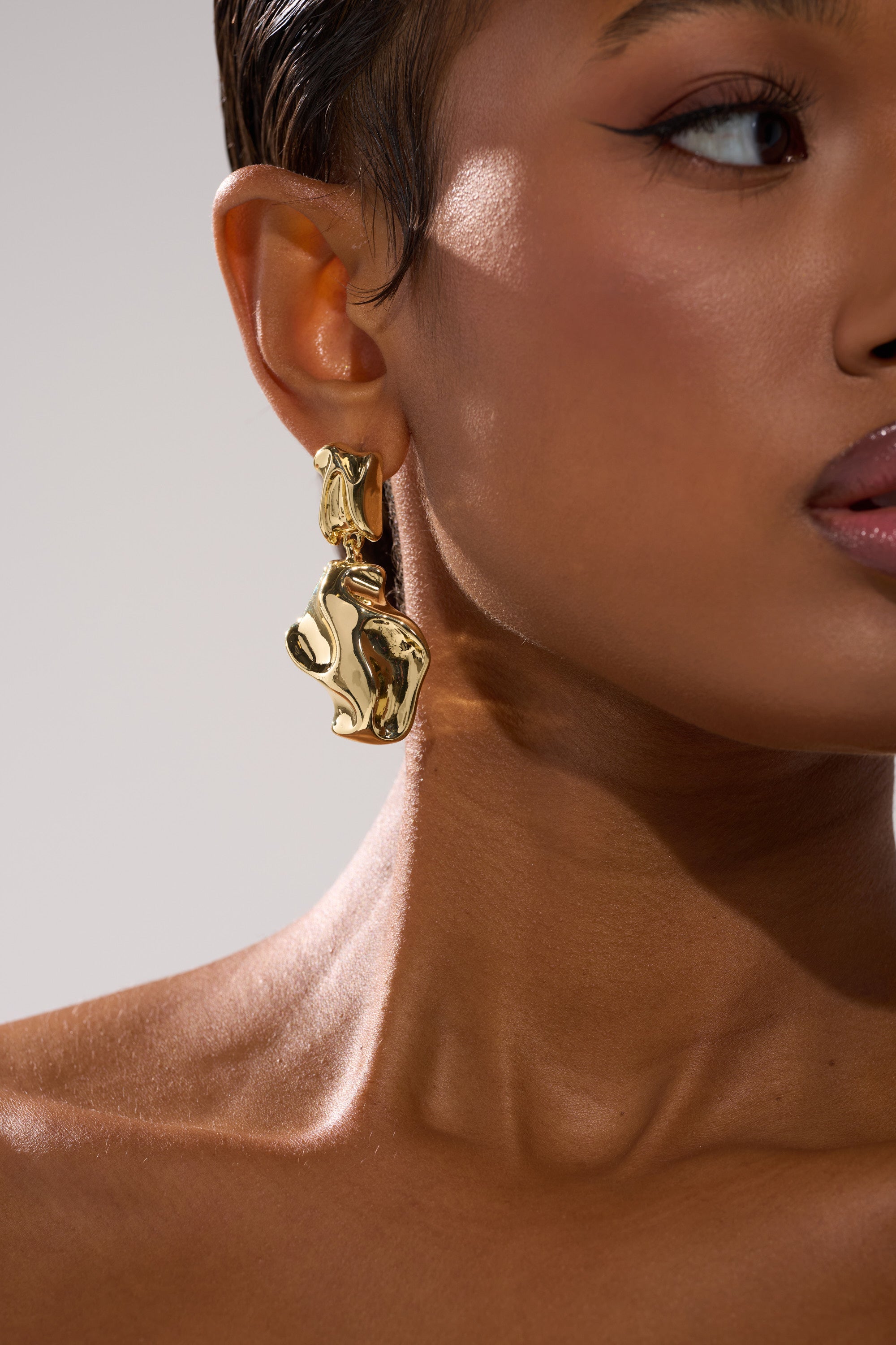 A close-up shows a person wearing the LEAVE ME ALONE EARRING in faux gold, highlighting their smooth skin, subtle eye makeup, and nude lips, with focus on the bold earring beside their face and neck.