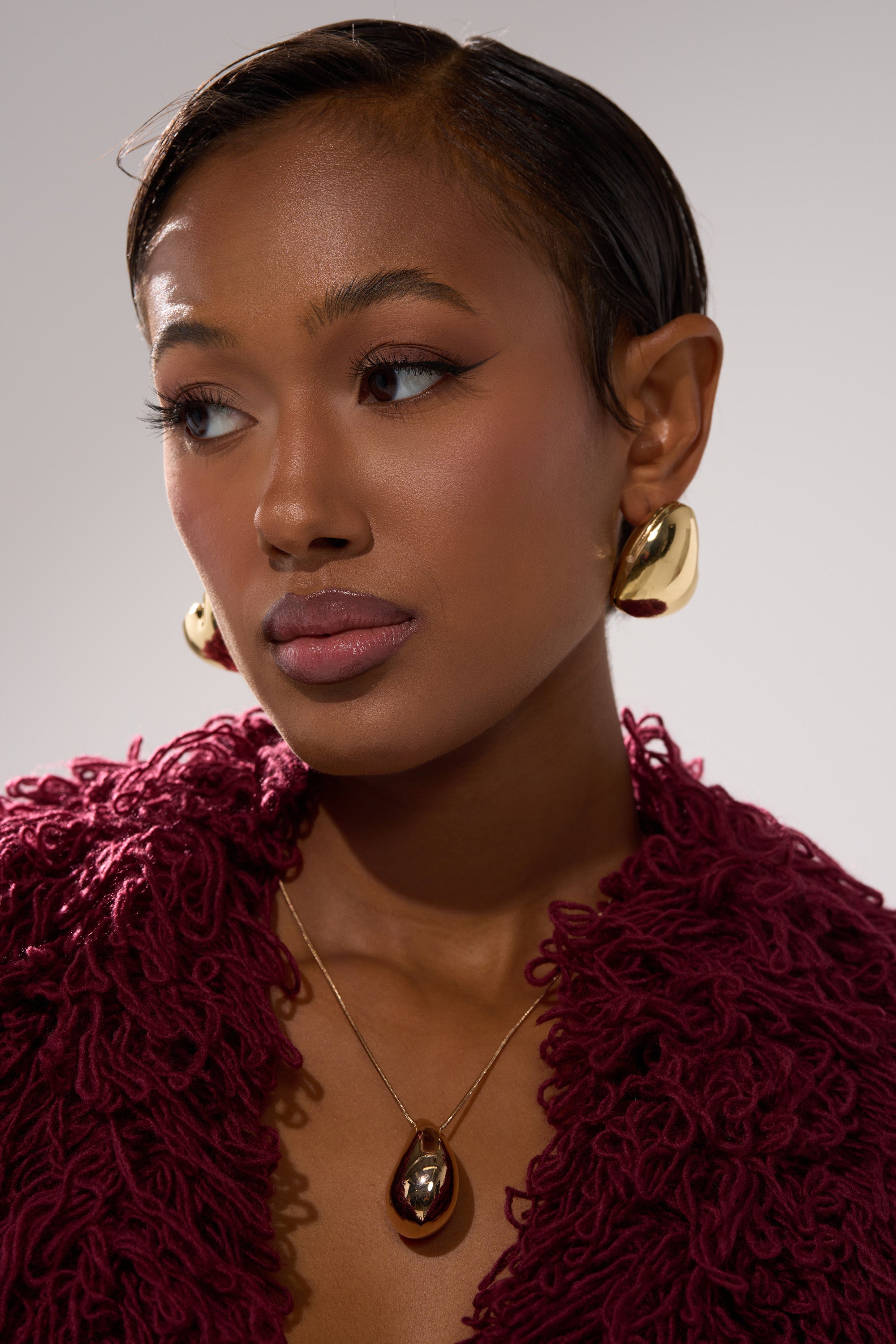 A woman with sleek, short hair wears the PUFFY HEART EARRING and a matching gold pendant necklace. She is dressed in a textured deep burgundy garment, gazing slightly to the side against a neutral background. 3