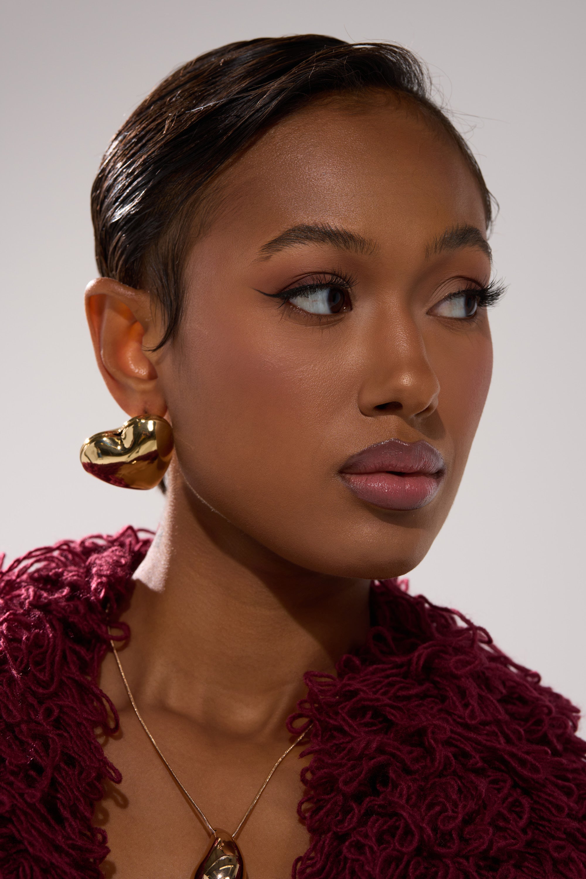 A woman with smooth brown skin and slicked-back hair wears a textured burgundy top and the PUFFY HEART EARRING. She looks to the side with a neutral expression. 4