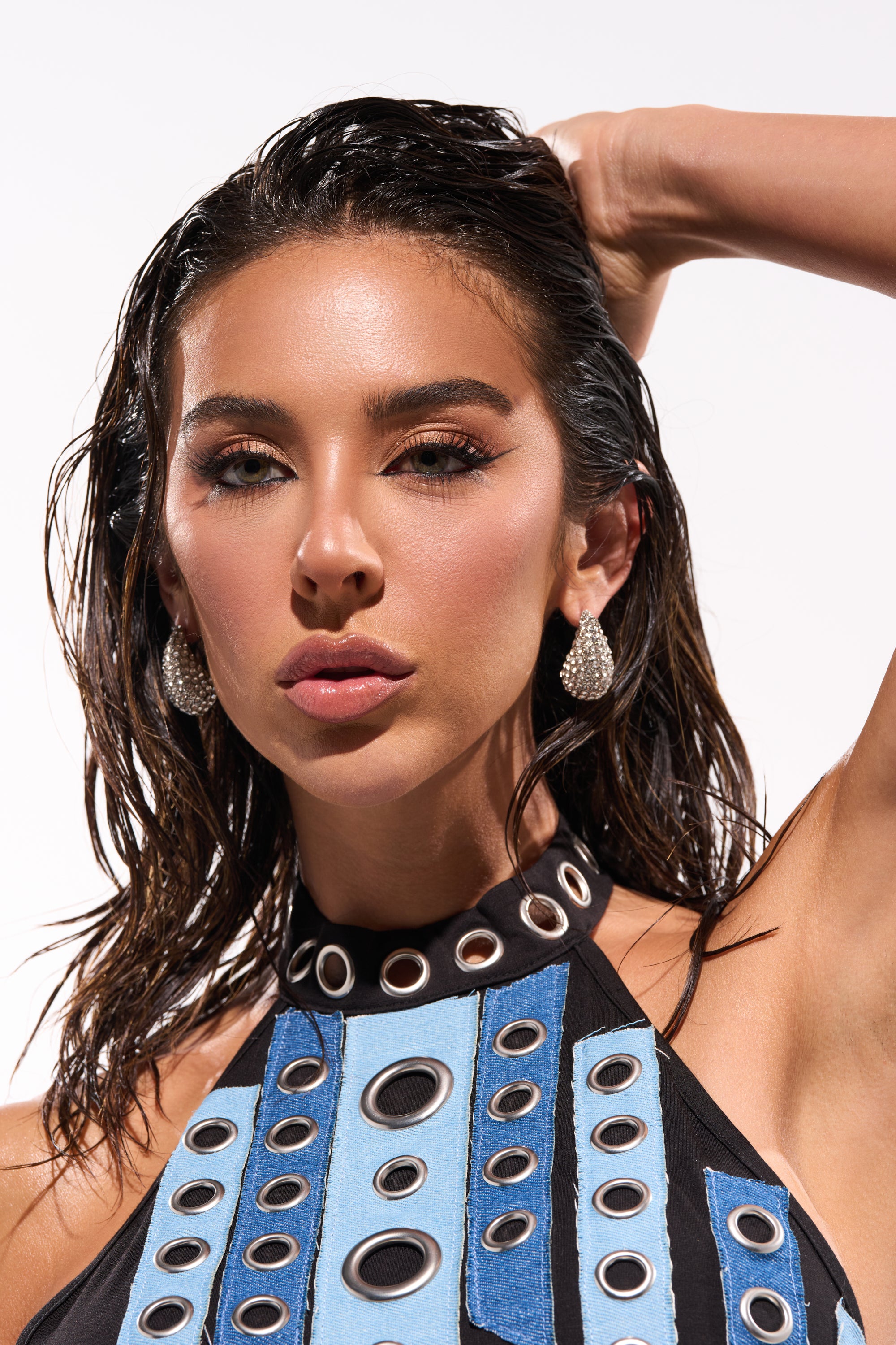 A woman with wet, dark hair poses with one arm raised behind her head, wearing the DROPS OF JUPITER EARRING IN SILVER and a sleeveless top with blue, black, and white circular-patterned straps. The background is plain and bright. 0