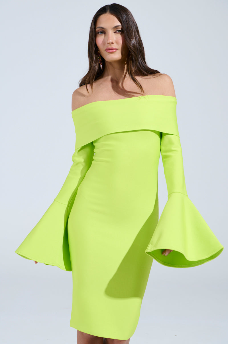 A woman in a "Work of Art" bandage midi dress in lime, featuring off-the-shoulder flared sleeves, stands against a plain backdrop. She has long dark hair and gazes slightly to the side with confidence.