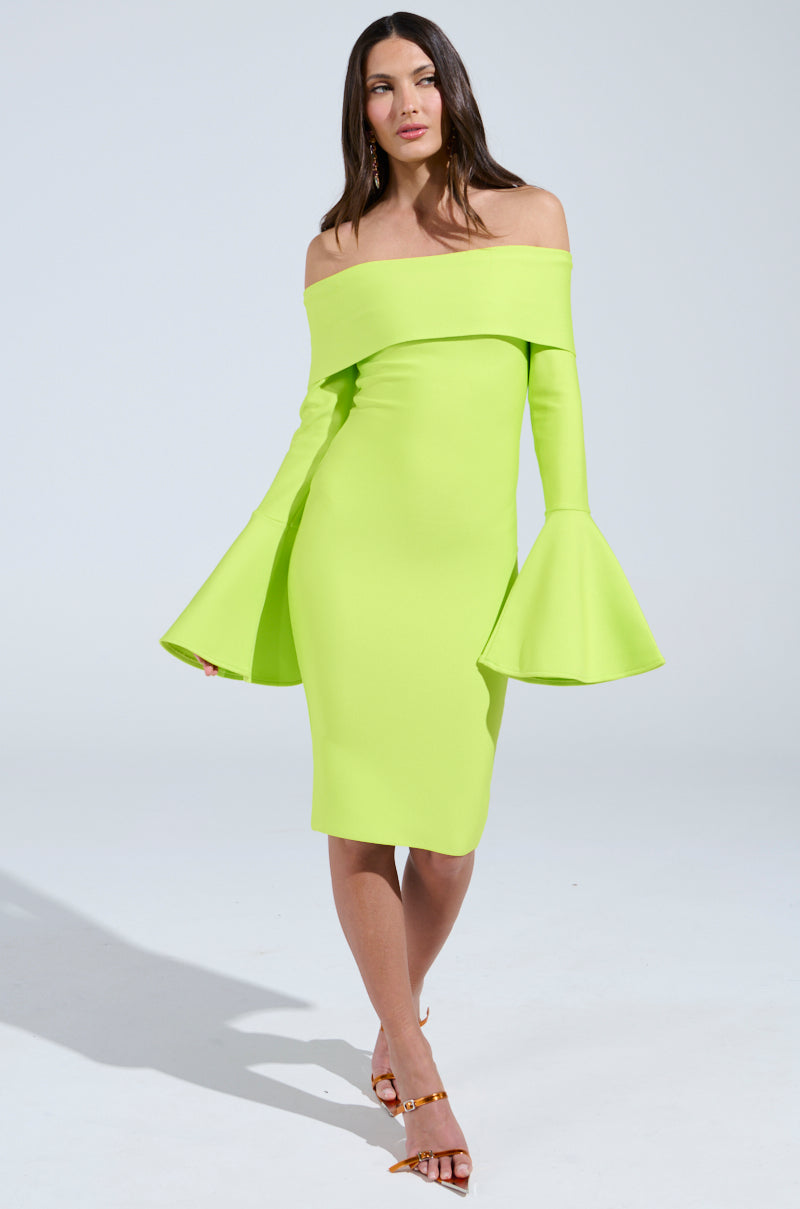 Wearing the "WORK OF ART BANDAGE MIDI DRESS IN LIME," a vibrant lime green off-the-shoulder midi with flared sleeves, a person pairs it with strappy brown sandals, all set against a plain white studio backdrop.