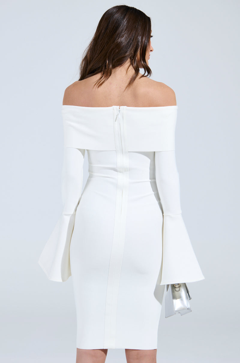 A woman in the WORK OF ART BANDAGE MIDI DRESS in white, with an off-the-shoulder and bodycon fit featuring flared cuffs, stands facing away, silver clutch in her left hand. The knee-length dress complements her long, wavy hair cascading elegantly down her back.