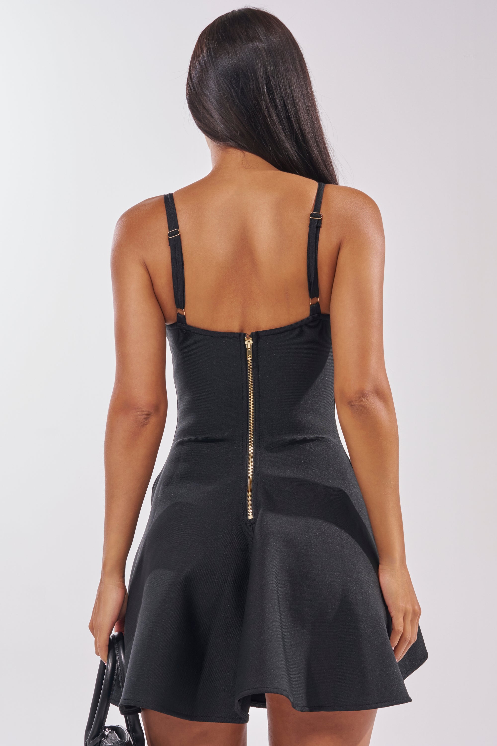 A woman with long straight hair, seen from behind, wears the ROXY BANDAGE MINI DRESS with adjustable tank straps and a visible gold zipper down the back, holding a black handbag.