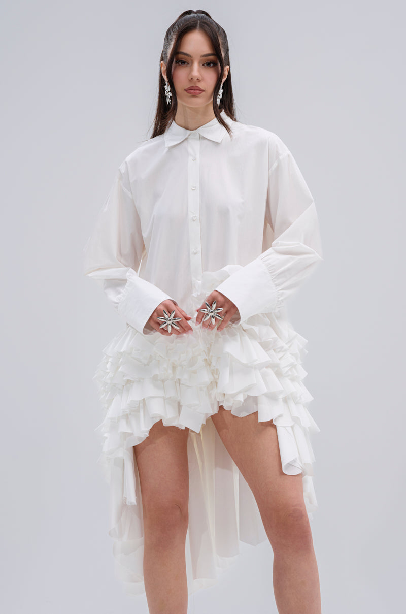 A woman wears the KIND OF A BIG DEAL POPLIN HIGH LOW DRESS IN WHITE, featuring a high-low hem, ruffled skirt layers, long sleeves, and a classic collar. She pairs it with large floral earrings and oversized star-shaped rings against a plain background.