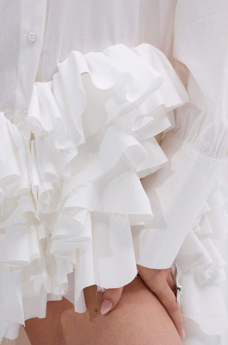 Close-up of a person’s hand by their thigh, wearing the KIND OF A BIG DEAL POPLIN HIGH LOW DRESS IN WHITE—a crisp, structured shirt dress with layered ruffled details for voluminous, textured folds.
