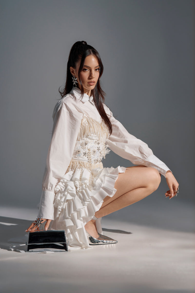 A woman with long dark hair kneels on one knee in a studio, wearing the KIND OF A BIG DEAL POPLIN HIGH LOW DRESS IN WHITE styled as a chic shirt dress. She accessorizes with silver earrings, silver shoes, and a small black purse against a gray background.