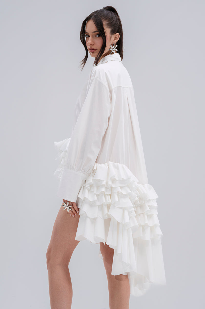 A woman poses sideways in the KIND OF A BIG DEAL POPLIN HIGH LOW DRESS IN WHITE, featuring long sleeves, dramatic layered ruffles, and a high-low hemline. She wears her long dark hair in a high ponytail, with bold star-shaped earrings and a matching ring.