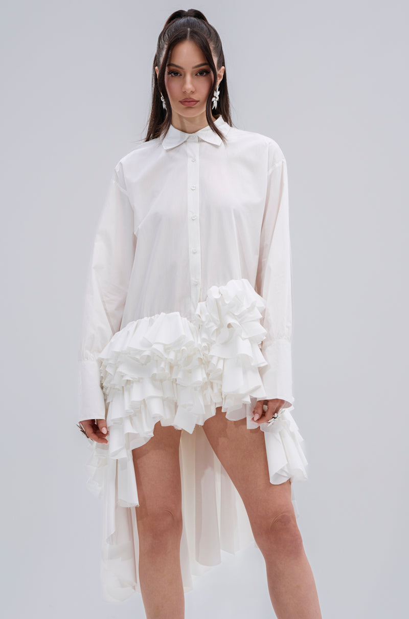 A woman models the KIND OF A BIG DEAL POPLIN HIGH LOW DRESS IN WHITE, featuring long sleeves, an asymmetrical high-low hemline, and ruffled front details, standing against a plain light gray background.