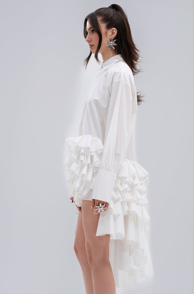 A woman stands in profile wearing the KIND OF A BIG DEAL POPLIN HIGH LOW DRESS IN WHITE, featuring dramatic ruffled layers at the hips and back. She accessorizes with star-shaped earrings and rings, her long dark hair styled in a high ponytail. The background is light grey.