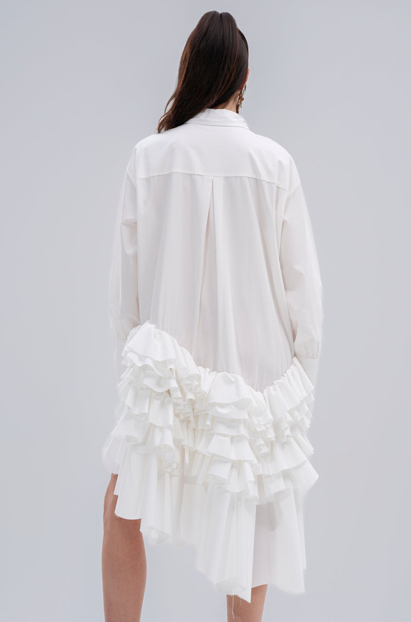 A person stands facing away, wearing the KIND OF A BIG DEAL POPLIN HIGH LOW DRESS IN WHITE, featuring long sleeves and layered ruffles on a loose high-low hem. The background is plain light gray.