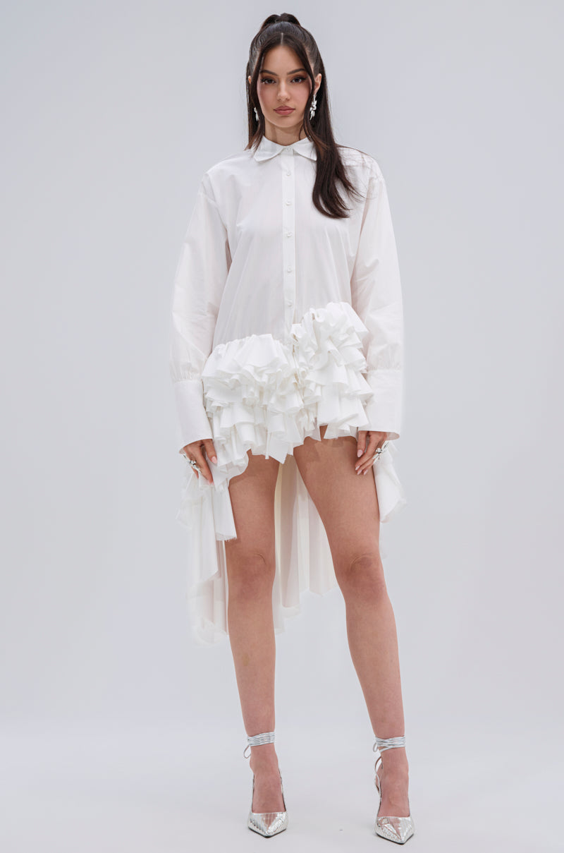 A woman stands against a plain background wearing the KIND OF A BIG DEAL POPLIN HIGH LOW DRESS IN WHITE with ruffled hip detailing, paired with silver high heels.