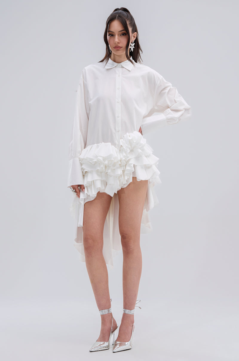 A woman stands against a plain background wearing the KIND OF A BIG DEAL POPLIN HIGH LOW DRESS IN WHITE, featuring a high-low hemline and ruffled hip details, paired with silver pointed-toe heels and statement earrings.