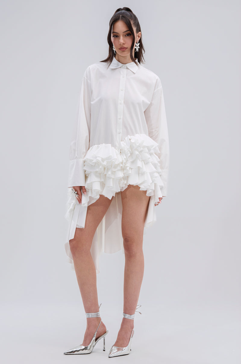 A woman models the KIND OF A BIG DEAL POPLIN HIGH LOW DRESS IN WHITE, featuring ruffled hip accents and a high-low hemline. She pairs it with metallic silver heels and statement earrings, posing against a plain light background.