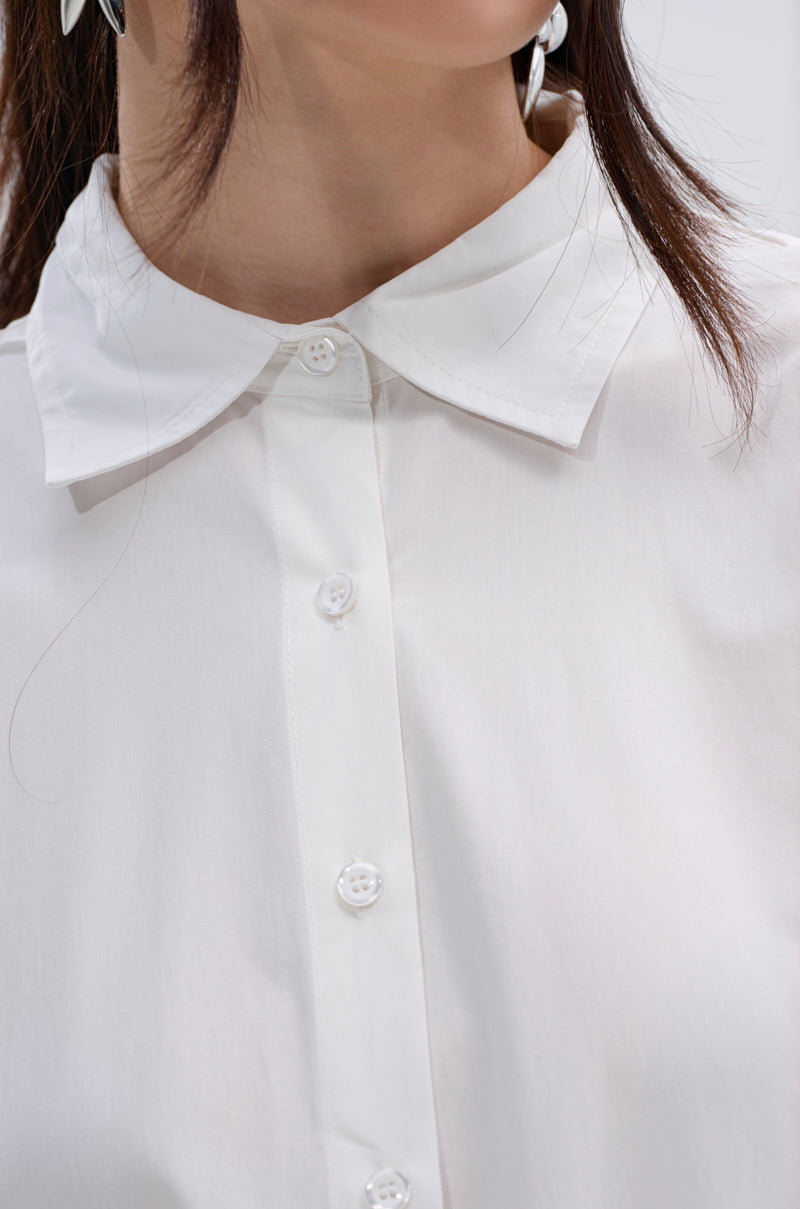 Close-up of a person wearing the KIND OF A BIG DEAL POPLIN HIGH LOW DRESS IN WHITE. Only the lower face, neck, and upper chest are visible. The dress features white buttons, and long dark hair is partially shown.