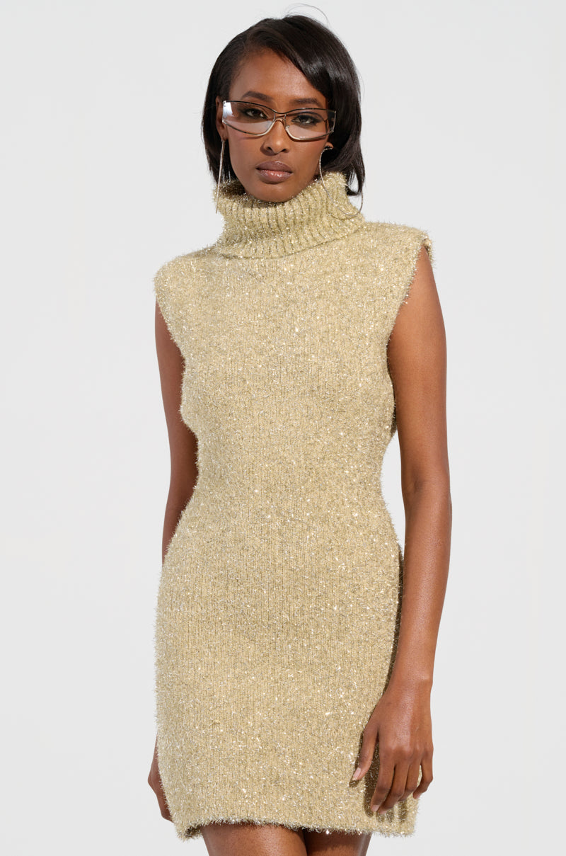 A woman strikes a pose in the FELT A SPARK KNIT GLITTER MINI DRESS, featuring a sleeveless, metallic gold and silver turtleneck design against a plain background. Her glasses catch the light, complementing her shoulder-length hair and adding a touch of sophistication to the glittering ensemble.