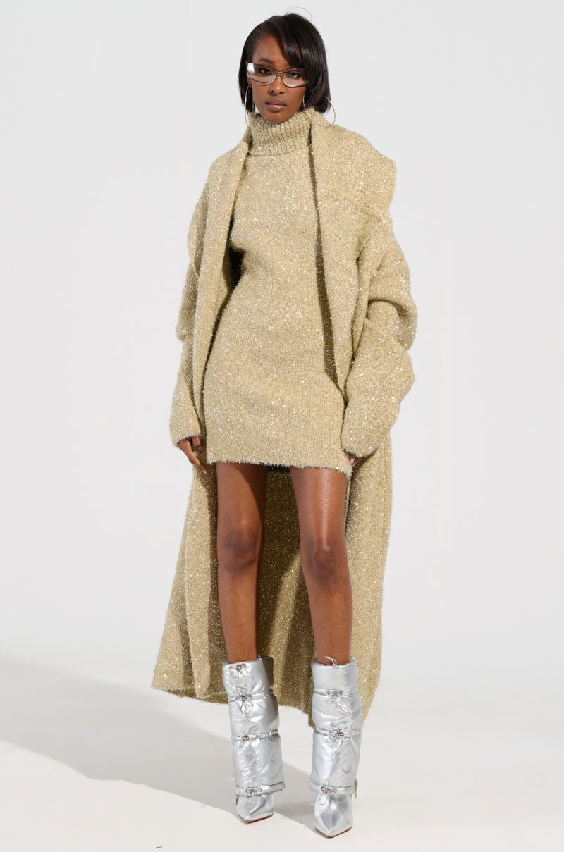A person exudes confidence in the FELT A SPARK KNIT GLITTER MINI DRESS, flawlessly complemented by silver high-heeled boots and dark glasses. The minimalist white background enhances their chic attire, emphasizing the sophisticated charm of their ensemble.