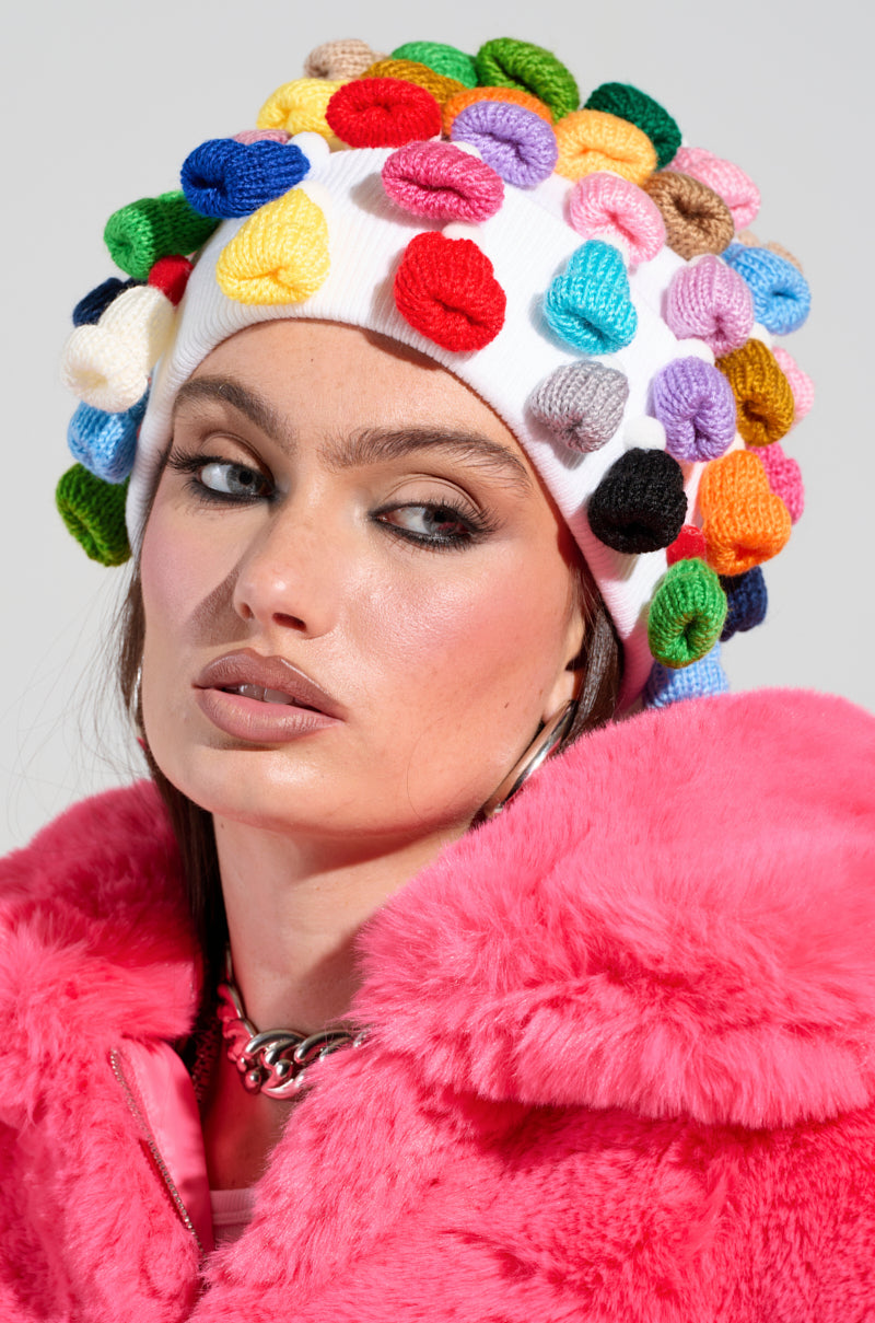 A person wearing the ME AND MY HATS BEANIE with vibrant pom-poms and a bright pink fur coat gazes to the side. They have long brown hair, bold eye makeup, and large hoop earrings against a neutral gray background.