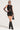 The woman wears a stylish GELINA MESH LACE UP MINI DRESS, featuring intricate patterns and a sleeveless design. She completes her look with long, semi-sheer gloves, large sunglasses, a small black handbag, and knee-high black boots. She poses against a plain light background.