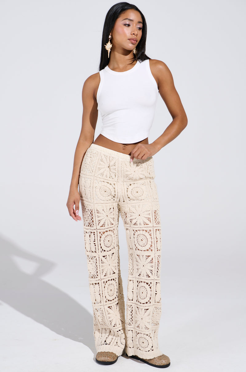 A woman exudes confidence in a white sleeveless crop top and CROCHET KNIT PANT with intricate square designs. Complemented by star-shaped earrings, her ensemble is the perfect high-rise fit for a summer slay against the plain white backdrop.