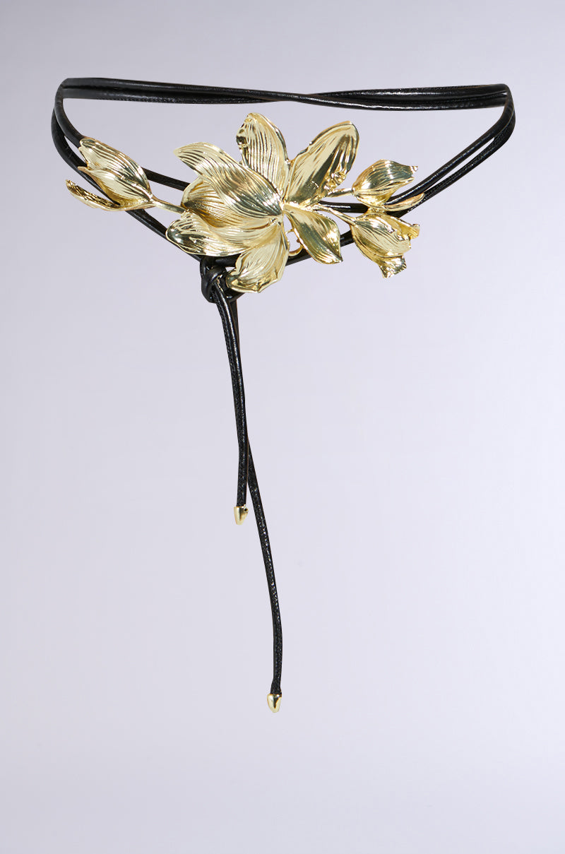 Introducing the PETAL POWER METAL DETAIL BELT: a black belt with an ornate gold floral design of large petals and small buds. Two thin cords hang at the center, ending with small gold caps. A striking accessory against a plain light gray background. 1