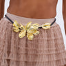 A person in a beige crop top and a tiered, ruffled skirt showcases the PETAL POWER METAL DETAIL BELT, featuring an intricate gold floral design. The focus is on the striking belt and detailed skirt.