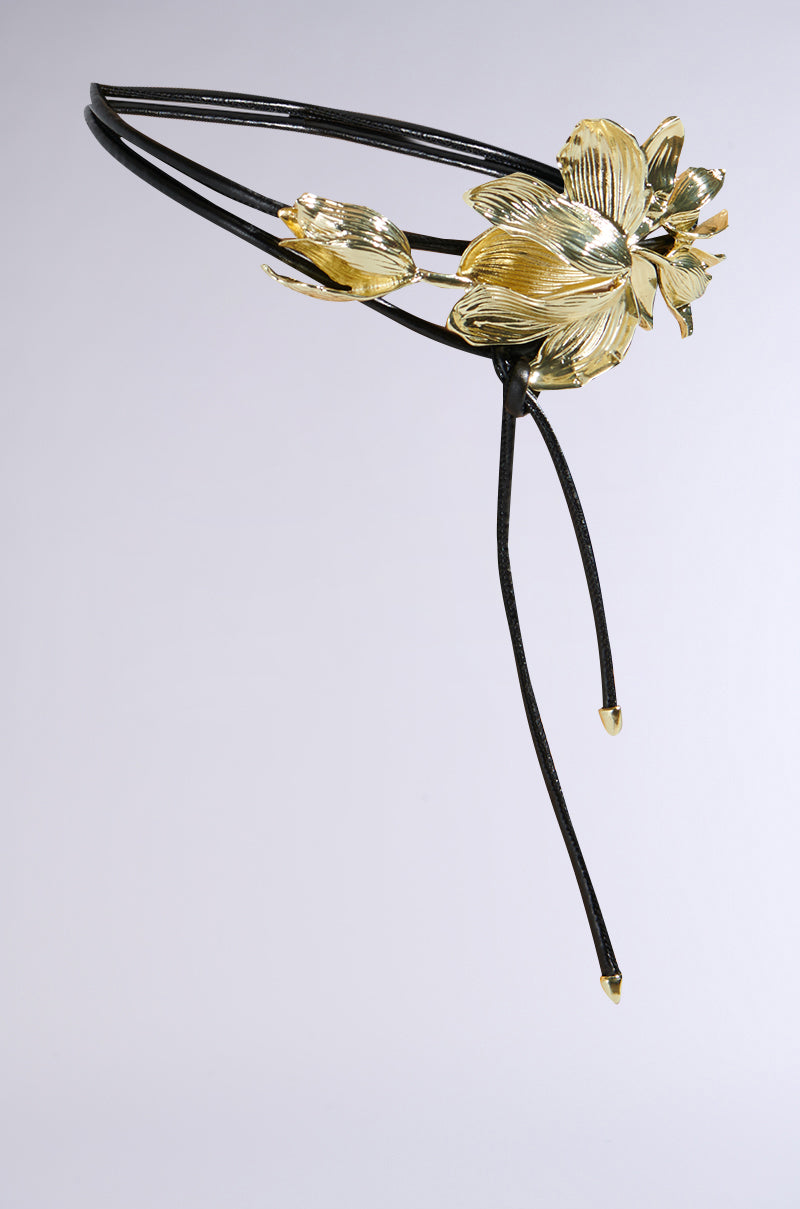 This statement accessory, the Petal Power Metal Detail Belt, features a large gold floral design with detailed petals and leaves. It includes two intersecting thin black bands at the floral centerpiece, finished with dangling ends and gold tips.