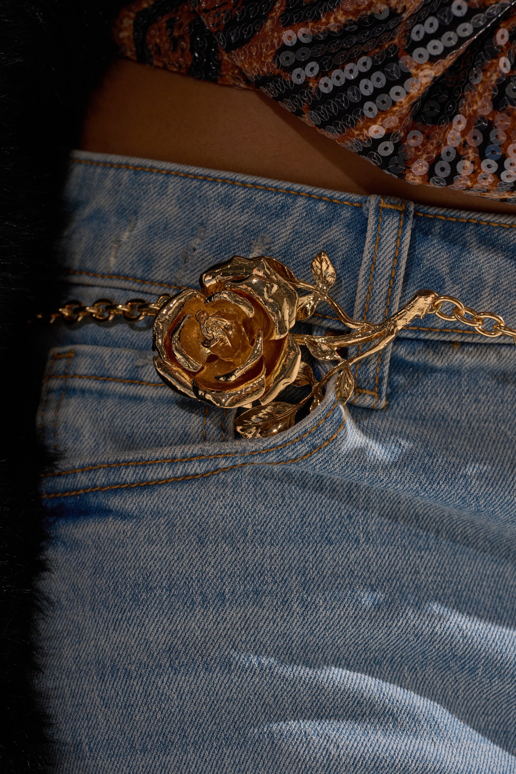 Close-up of blue denim jeans paired with the LIKE A ROSE CHAIN BELT, featuring a gold rose-shaped buckle. A sequined brown, black, and silver top adds shimmer. Cropped to highlight the stylish waist detail.