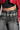 A person is styled in a black top and red leather jacket, paired with high-waisted black jeans embellished with silver heart decorations and shiny beads along the waistband. The look is completed with the chic HOLD MY HEART METAL CHAIN BELT. Only the midsection and partial arms are visible.